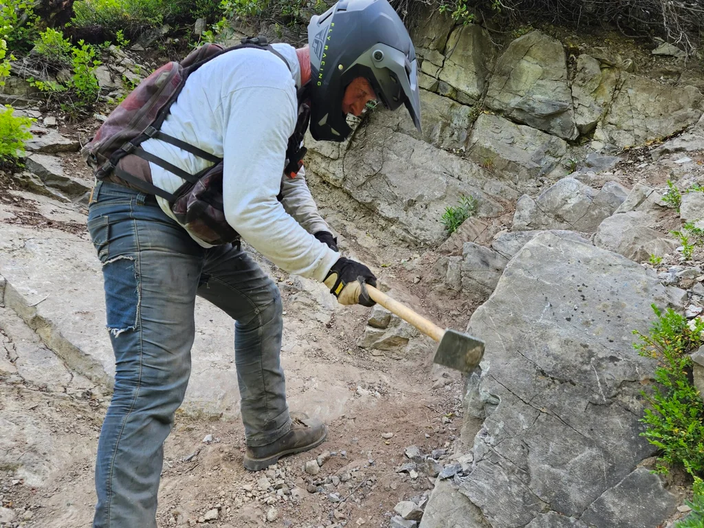 A volunteer in a motorcycle helmet breaking rock out of the trail with a hand tool.